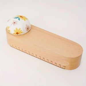 May include: A wooden seamstress's clapper with a floral pin cushion. The clapper has a ruler etched along its side, measuring up to 15 cm (5.9 inches). The pin cushion is white with yellow and pink floral patterns.