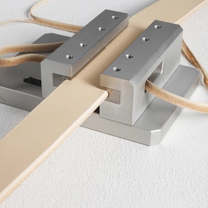 May include: A metal leatherworking tool with a light tan leather strip being fed through it. The tool has a silver finish and is designed for precise cutting or shaping of leather. The leather strip is approximately 2.5 cm wide.