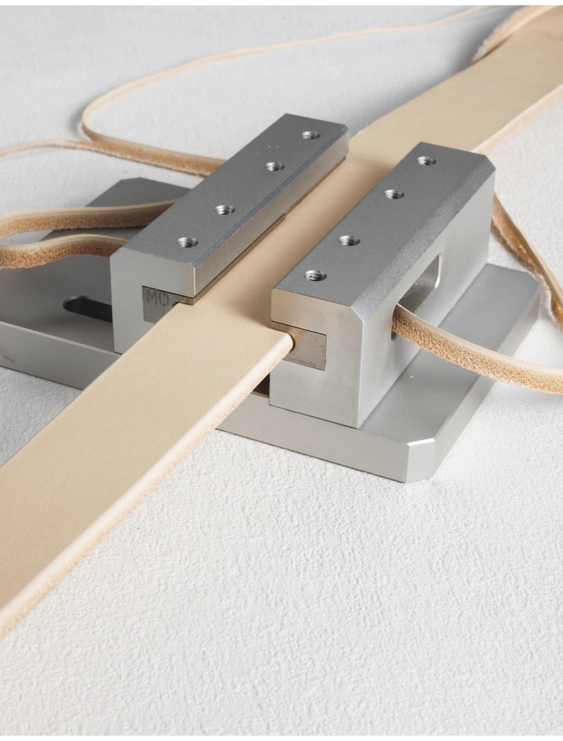 May include: A metal leatherworking tool with a light tan leather strip being fed through it. The tool has a silver finish and is designed for precise cutting or shaping of leather. The leather strip is approximately 2.5 cm wide.