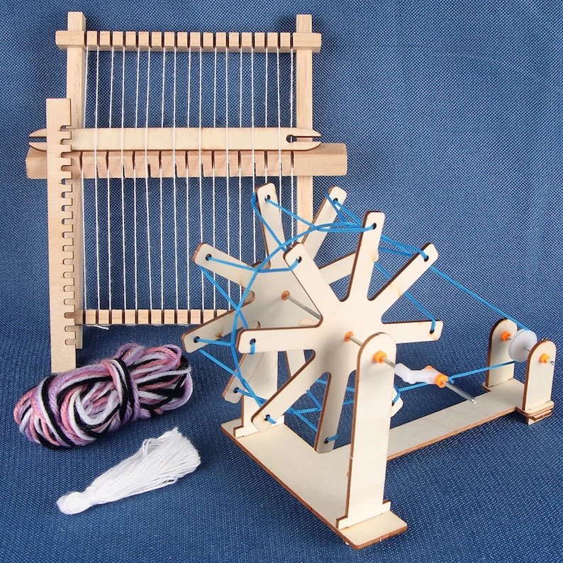 May include: A wooden loom with a spinning wheel and yarn. The loom is made of natural wood and has a blue string threaded through it. The spinning wheel is also made of wood and has a blue string threaded through it. The yarn is pink, white, and black.