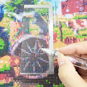 Puede incluir: Primer plano de una pintura de diamantes en progreso. Una guía de metal plateado se coloca sobre el lienzo, con un bolígrafo transparente aplicando una gema. El lienzo está lleno de gemas coloridas y brillantes, creando una imagen tipo mosaico.