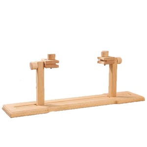 Puede incluir: Un soporte de bastidor de bordado de madera con una base rectangular y dos soportes verticales ajustables. El soporte está hecho de madera de color claro y está diseñado para sujetar un bastidor de bordado en su lugar para coser.