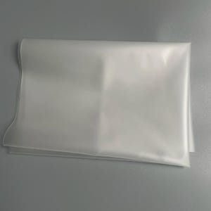 Op de afbeelding: Een gevouwen, doorschijnend wit vel materiaal, mogelijk plastic of stof, ligt op een grijze ondergrond. Het vel heeft een gladde textuur en is netjes gevouwen, met zichtbare vouwen en randen.