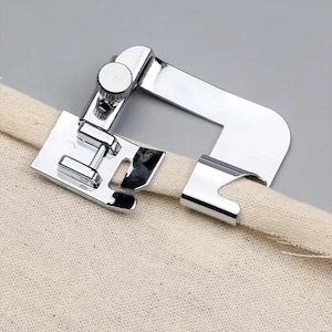 May include: A silver-coloured metal sewing machine presser foot attachment designed for creating hems. The device features a flat, U-shaped guide arm and a small adjustable screw mechanism, currently positioned over the edge of a piece of beige fabric.