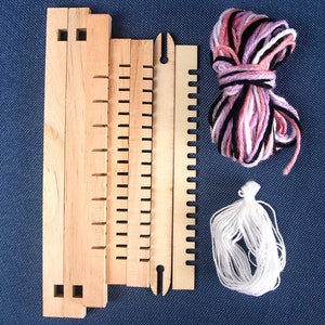 May include: A wooden loom kit with four pieces of wood, a shuttle, and two balls of yarn. One ball of yarn is pink, white, and black, and the other ball is white.