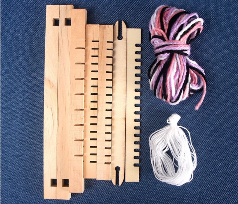 May include: A wooden loom kit with four pieces of wood, a shuttle, and two balls of yarn. One ball of yarn is pink, white, and black, and the other ball is white.