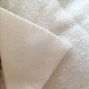 Op de afbeelding: Close-up van witte, donzige stof. Het materiaal lijkt zacht en getextureerd, met een enigszins ongelijk oppervlak. De stof is opgevouwen en onthult meerdere lagen en een gewatteerd patroon.