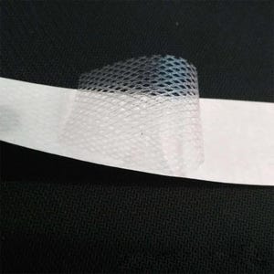 Può includere: Una striscia di nastro adesivo bianco con una sezione di rete traslucida a maglia fine attaccata alla superficie. La rete ha un distinto motivo a forma di diamante ed è parzialmente sollevata dal supporto bianco, mostrando una consistenza semitrasparente.