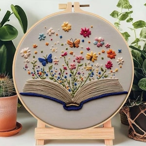 以下が含まれることがあります： 開いた本からカラフルな花と蝶が飛び出す刺繍の作品。青、ピンク、黄色、オレンジなどの色が使われ、薄いグレーの背景に施されています。木製のフレームは、小さな木製のイーゼルに飾られています。