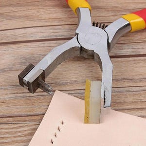 May include: A close-up of a silver leather punch tool with red and yellow handles. The tool is being used to punch holes in a piece of light brown leather. The tool has a small, square punch head and a long, thin handle.