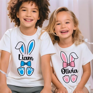 May include: Two white t-shirts with bunny ears, one with blue ears and a blue bow tie with the name "Liam" and the other with pink ears and a pink bow with the name "Olivia".