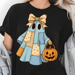 以下が含まれることがあります： 黒いTシャツ。パッチワークデザインの青、オレンジ、黄色の漫画の幽霊が描かれています。幽霊はジャック・オー・ランタンのバケツを持っています。大きなリボンが幽霊の頭を飾っています。「PNG - INSTANT DOWNLOAD」の文字が下にあります。