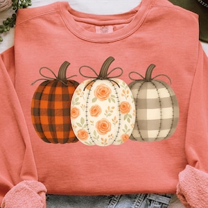 May include: A coral-colored sweatshirt featuring three pumpkin designs. The pumpkins have different patterns: a plaid, a floral, and a checkered design. The text "PNG - INSTANT DOWNLOAD" is at the bottom of the image.