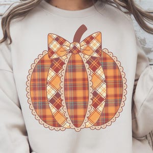 Könnte beinhalten: Ein beiges Sweatshirt mit einem Kürbisdesign. Der Kürbis ist orange, rot und gelb kariert mit einem braunen Stiel und einer karierten Schleife. Das Design hat einen gewellten Rand. Der Text "PNG - INSTANT DOWNLOAD" steht unten.