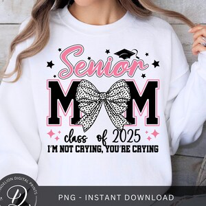 May include: White sweatshirt featuring the text "Senior Mom" in pink script and black block letters. A bow graphic is centered between the letters "M". The text "class of 2025" and "I'M NOT CRYING, YOU'RE CRYING" are also displayed.