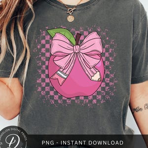 May include: Dark gray t-shirt featuring a pink apple design with a large bow, pencil, and green leaf. The apple is set against a pink and gray checkered background. The text "PNG - INSTANT DOWNLOAD" is at the bottom.