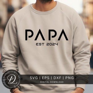 Peut inclure: Un sweat-shirt beige avec le texte "PAPA EST 2024" en noir. Le texte est en gras, police sans empattement. Sous le texte, il y a un logo avec le texte "DANDELION CUT-READY SVG" et une lettre stylisée "D". Le texte "SVG | EPS | DXF | PNG DIGITAL DOWNLOAD" est également visible.