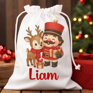 Puede incluir: Bolsa de regalo blanca con cordón con una ilustración de dibujos animados de un reno con una bufanda roja y un cascanueces sosteniendo un regalo. El nombre "Liam" está impreso en escritura roja. La bolsa está decorada con elementos navideños.