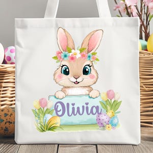 Può includere: Borsa tote in tela bianca con un coniglio dei cartoni animati che indossa una corona di fiori. Il coniglio tiene un cartello che dice "Olivia". La borsa è decorata con uova di Pasqua color pastello e fiori.