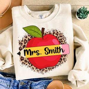 May include: A cream-colored sweatshirt with a red apple and a yellow pencil with the text "Mrs. Smith" on it. The apple is on a brown leopard print background.