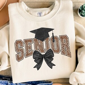 May include: A cream-coloured sweatshirt featuring the word "SENIOR" in a leopard print font, a black graduation cap, and a black bow. This casual garment is ideal for celebrating a graduation.