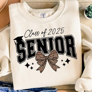 May include: A cream-coloured sweatshirt featuring the text "Class of 2025 SENIOR" in black and white. The design includes a graduation cap and a leopard print bow. Four black star shapes are also included.