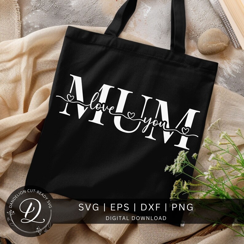 May include: A black tote bag with the text "MUM love you" in white with small hearts on either side of the text. The text is in a cursive font. The bag is on a beige surface with white flowers.