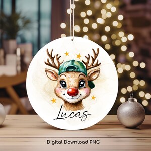May include: A round Christmas ornament featuring a cartoon reindeer wearing a green cap and a red nose. The ornament has the name "Lucas" written in cursive and is decorated with gold stars. The words "Digital Download PNG" are at the bottom.