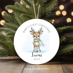 Può includere: Ornamento rotondo bianco con un'illustrazione ad acquerello di un cervo bambino che indossa una sciarpa verde. L'ornamento dice "Baby's First Christmas" e "Lucas - 2024 -" in testo nero.