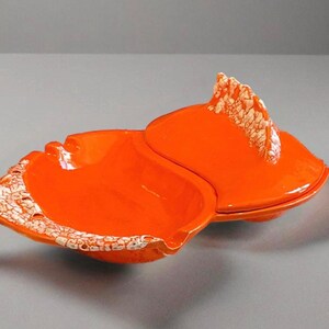 May include: Bright orange ceramic dish with a lid, resembling a seashell. The edges of the dish and lid feature a textured, off-white pattern. The dish is open, revealing its interior.