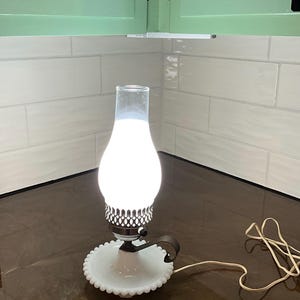 May include: A vintage-style electric lamp with a white, pear-shaped glass shade. The lamp has a white base with a scalloped edge and a dark metal handle. The lamp is lit, emitting a soft glow. A white cord extends from the base.