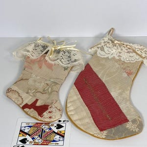 May include: Two handmade Christmas stockings made from patchwork fabric with lace trim. The stockings are beige, red, and cream with floral patterns. The larger stocking has a red stripe.