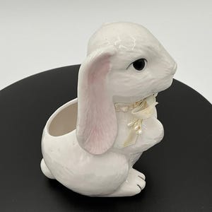 May include: White ceramic bunny planter with a pink ear and a white ribbon with pearl accents. The planter has a small opening at the top for planting.