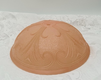 May include: A peach-colored, dome-shaped lampshade with an etched floral design. The glass shade has a textured surface and a decorative, swirling pattern. The lampshade has a small hole at the top and a rounded edge.