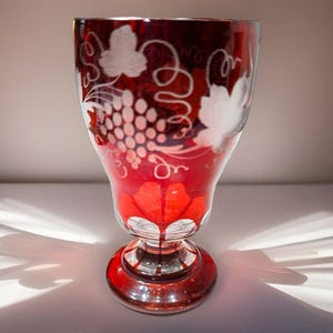 May include: A ruby red wine glass with a detailed etched grape and vine design. The glass features a round base and a flared top. The etched design is white, creating a contrast against the red glass. The glass is approximately 6 inches tall.