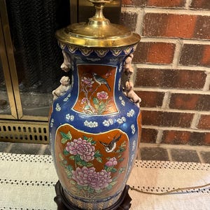 May include: A vintage ceramic table lamp with a brass top and base. The lamp features a blue and orange vase with floral and bird designs. The vase is mounted on a dark wooden base. The lamp is near a brick wall.