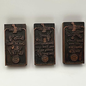 May include: Three rectangular, vintage printing blocks with copper-colored borders and dark gray centers. Each block features a different design and text, including a car, people, and various symbols. The text is difficult to read.