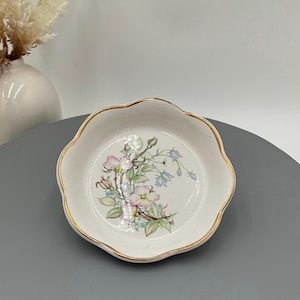 May include: A small, white, scalloped-edge dish with a gold rim. The dish features a floral design with pink and blue flowers and green leaves. The dish is likely made of porcelain or ceramic.