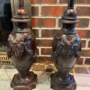 May include: Two bronze-colored table lamps with ornate detailing. Each lamp features a base with a square design, a central urn-shaped body with a goat head motif, and a dark brown neck. The lamps are topped with a light-colored lamp shade fitting.