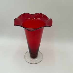 May include: A tall, ruby red glass vase with a flared, ruffled rim. The vase has a slender, conical body and a clear, circular base. The glass appears smooth and glossy, reflecting light. The vase is likely for decorative purposes.