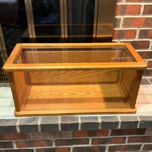 May include: A rectangular wooden display case with a glass top and sides. The case is made of light-colored wood and has a simple, classic design. The interior is empty, ready for showcasing collectibles or other items.