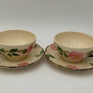 May include: Two cream-colored ceramic teacups and matching saucers featuring a hand-painted floral pattern of pink flowers and green leaves. Each cup has a dark brown, textured handle shaped like a twig, and the saucers have scalloped edges.