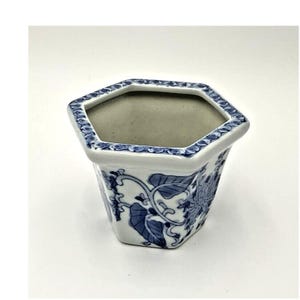 May include: A white ceramic hexagonal planter with a blue and white floral design. The planter has a wide opening and a slightly tapered base.