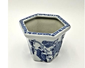 May include: A white ceramic hexagonal planter with a blue and white floral design. The planter has a wide opening and a slightly tapered base.
