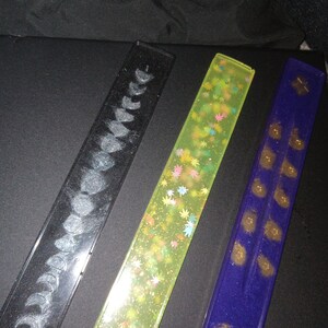 May include: Three long, rectangular incense holders. The first is black with a silver glitter design of crescent moons. The second is light green with a gold glitter design of leaves. The third is dark blue with a gold glitter design of dots.