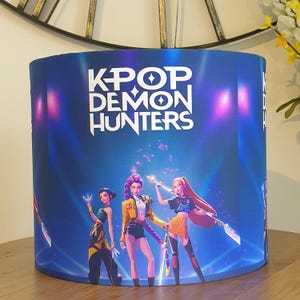 May include: A cylindrical lampshade with a blue backdrop, displaying the white text "KPOP DEMON HUNTERS". The design showcases three animated female figures in dynamic poses, wearing vibrant outfits. The theme is fantasy and pop culture.