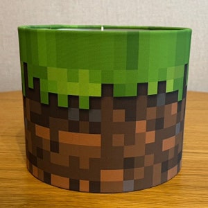 May include: A cylindrical lampshade with a green grass block and brown dirt block pattern, inspired by the popular video game Minecraft.