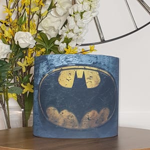 May include: A cylindrical container with a blue and gold Batman logo. The container has a distressed, textured finish. The Batman symbol is gold against a dark blue backdrop. The container sits on a wooden surface. The container is approximately 15 cm tall.