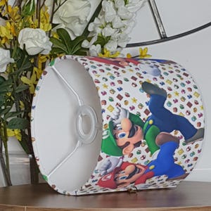 May include: A cylindrical lampshade featuring a colorful pattern of cartoon characters, including Mario and Luigi, on a white background. The design includes various game-related icons. The lampshade has a white interior and a metal frame.