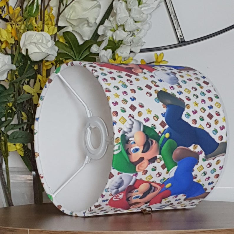 May include: A cylindrical lampshade featuring a colorful pattern of cartoon characters, including Mario and Luigi, on a white background. The design includes various game-related icons. The lampshade has a white interior and a metal frame.